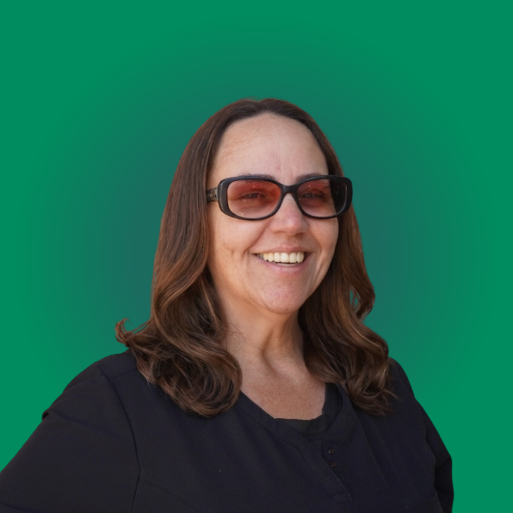 A smiling female dental team member with shoulder-length wavy brown hair, wearing black glasses and a black medical scrub top, posed against a solid green studio backdrop.