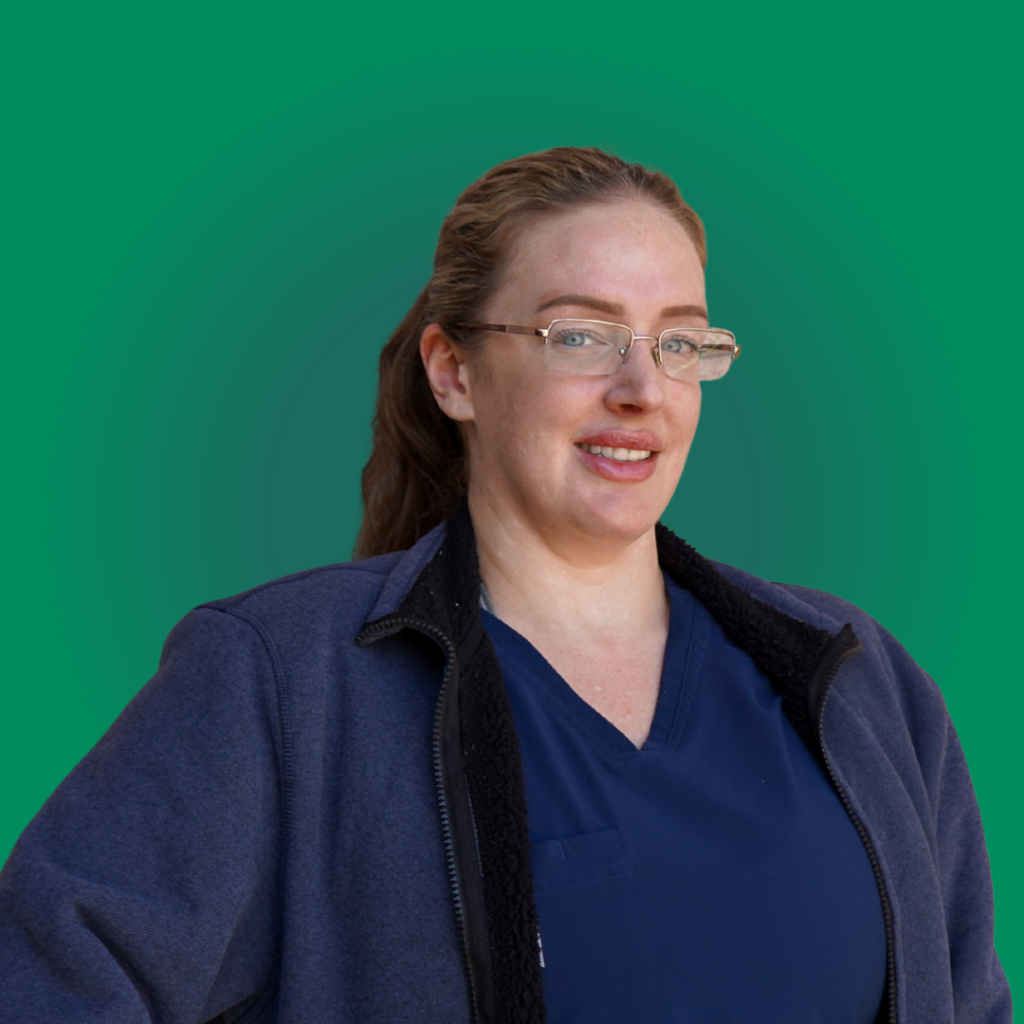 A smiling dental team member with glasses and a blue jacket over navy scrubs, pictured against a green background for Dr. Shirley Zhao's dental office in Pleasanton.