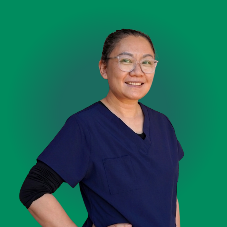 Dr. Shirley Zhao DDS, a female dentist in Pleasanton wearing clear glasses and navy blue scrubs, smiling and looking to the side against a solid green background.