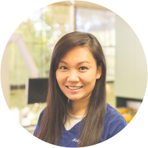 A circular cropped portrait of a smiling dental team member with long dark hair, wearing blue scrubs in front of a bright clinic window at Dr. Shirley Zhao's office in Pleasanton, CA.