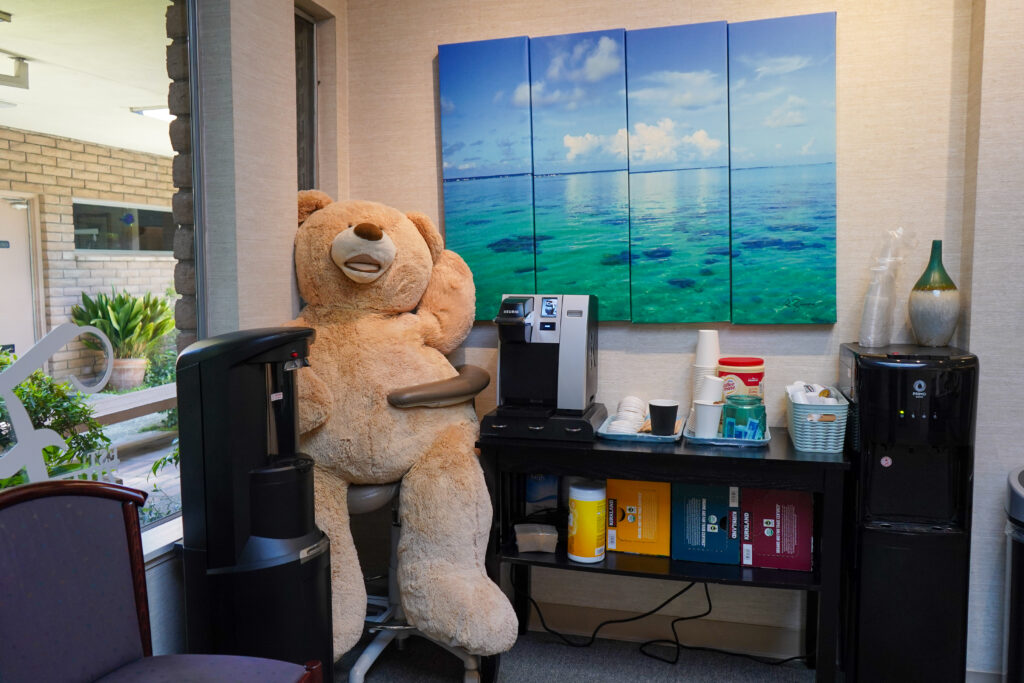 A cozy office beverage station featuring a coffee maker, water dispenser, and a large plush teddy bear sitting in a chair next to a tropical ocean wall art panel.